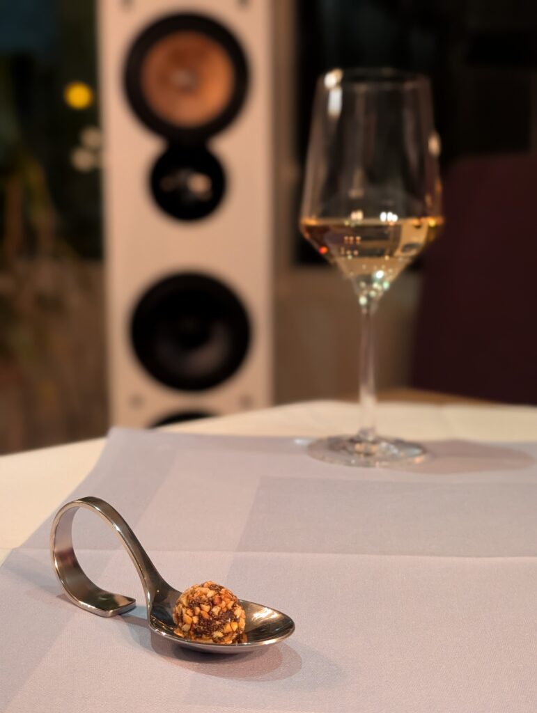 Nahaufnahme eines silbernen Degustationslöffels mit einer Nuss-Praline, im unscharfen Hintergrund sind ein weißer High-End-Lautsprecher und ein Weinglas zu sehen.